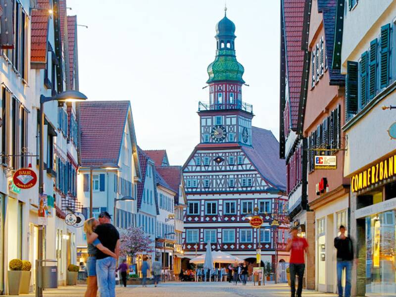 Eine Fußgängerzone in einer beleuchteten, dämmrigen Stadt. Geradeaus ist die Kirche und rechts sowie links sind Ladengeschäfte. In der Fußgängerzone sind ein paar vereinzelte Menschen unterwegs. 