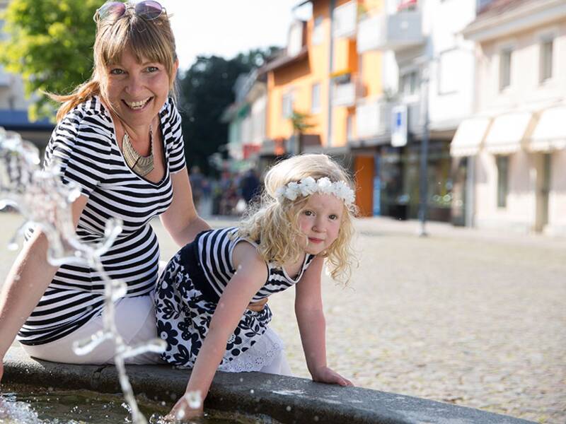 Eine Mutter mit ihrer kleinen Tochter, sie sitzen auf dem Rand eines Brunnens und halten ihre Hände in das Wasser. Im Hintergrund sind Ladengeschäfte der Stadt. 