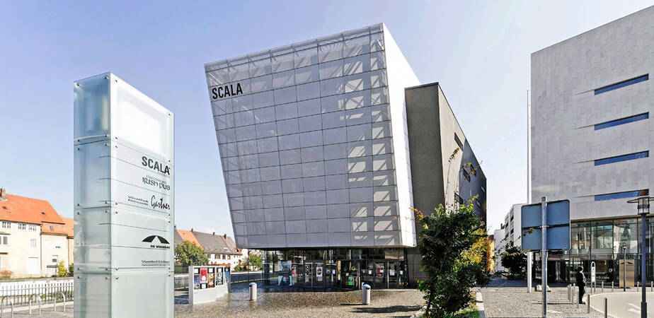 Großes, verglastes Gebäude auf einem Marktplatz. Rechts daneben ein weiteres großes Gebäude. 