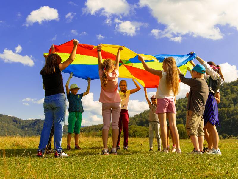 Mehrere Kinder stehen in einem Kreis und halten ihre Hände nach oben. Sie halten alle zusammen ein großes, buntes, ausgebreitetes Tuch in ihren Händen. Sie schauen sich unter dem Tuch durch an. 