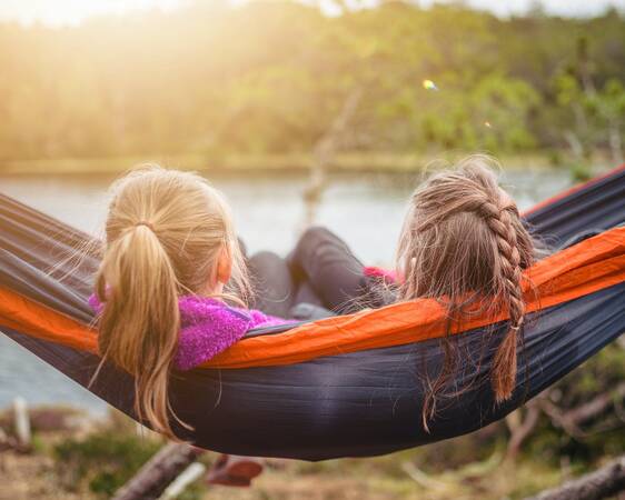 Eine Hängematte und im Hintergrund ein See. In der Hängematte liegen schräg zwei Frauen drin. Die beiden Köpfe sind von hinten drauf. Sie schauen in Richtung des Sees. 