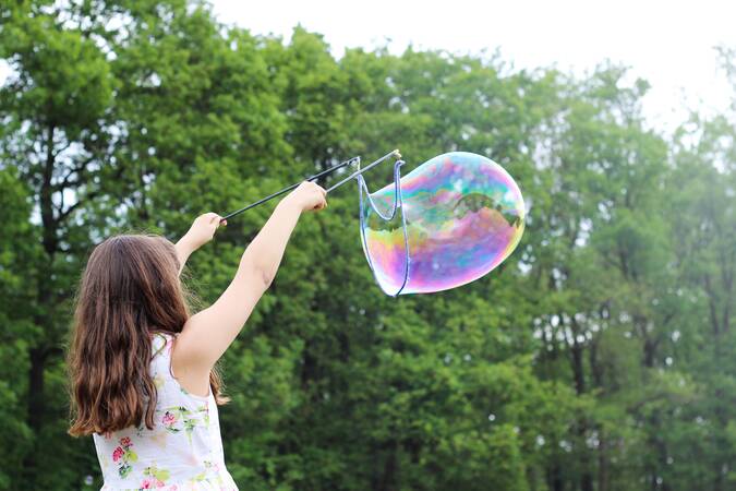 Ein  Kleides Mädchen macht riesige Seifenblasen mit einem extra Spielzeug dafür. Im Hintergrund stehen viele Bäume. Das Mädchen wurde von hinten fotografiert. 