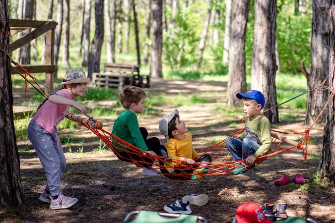 Drei kleine Jungs sitzen auf einer Netz-Hängematte im Wald zwischen vielen Bäumen. Ein Mädchen steht links daneben und hält sich an der Hängematte. 