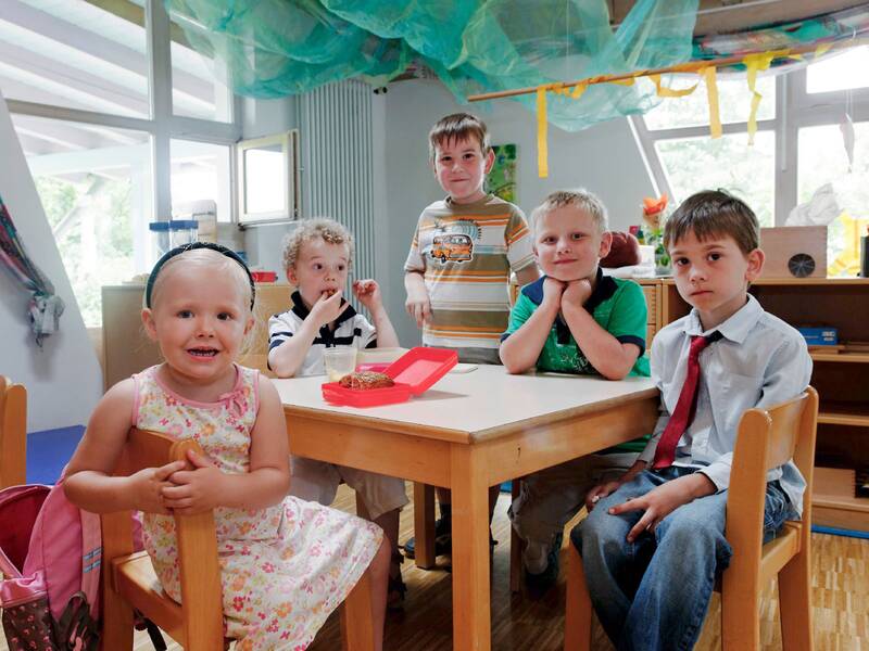 Fünf Kinder sitzen und stehen um einen kleinen Tisch herum. Einer ist gerade am Essen. Vier Kinder schauen in die Kamera.