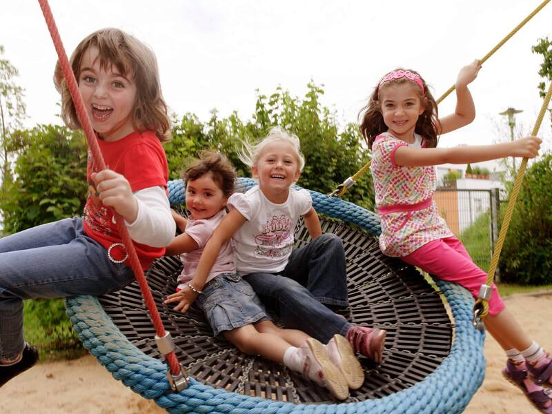 4 Kinder sitzen auf einer runden Schaukel mit einem Netz. Alle 4 Mädchen schauen in die Kamera und lachen.