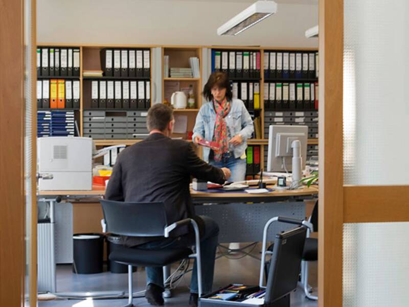 Auf dem Bild wurde das Büro in einem Rathaus durch den Türrahmen fotografiert. es steht ein Schreibtisch mitten im aum. Davor sitzt ein Mann auf einem Stuhl, der mit dem Rücken zur Kamera sitzt. Hinter dem Tisch, zur Kamera schauend steht eine Frau die sich mit dem sitzenden Mann unterhält. 