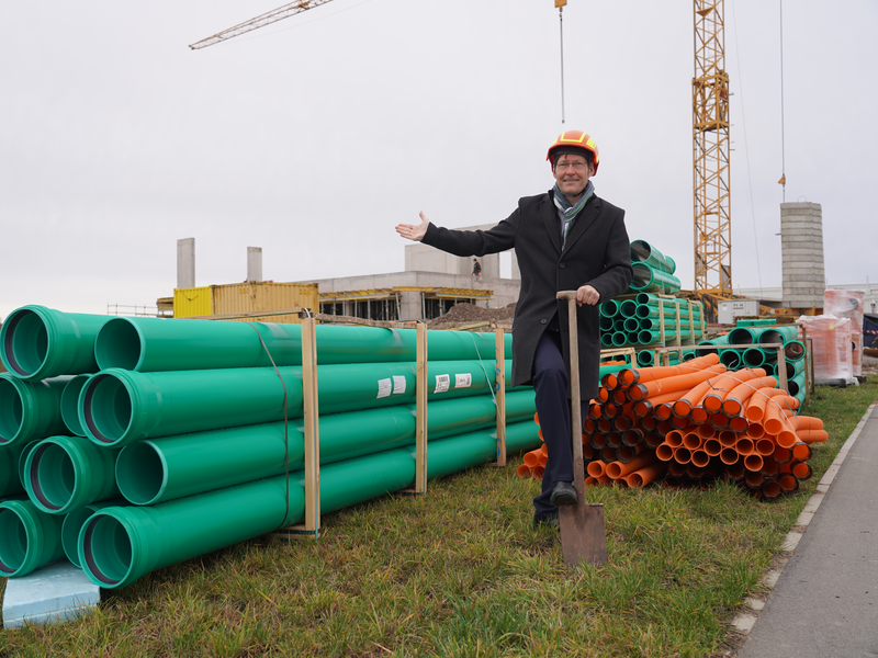 Ein Mann in einem Anzug und Bauhelm steht auf einer Baustelle neben großen grünen und orangen Rohren, während er lächelnd eine Schaufel hält.