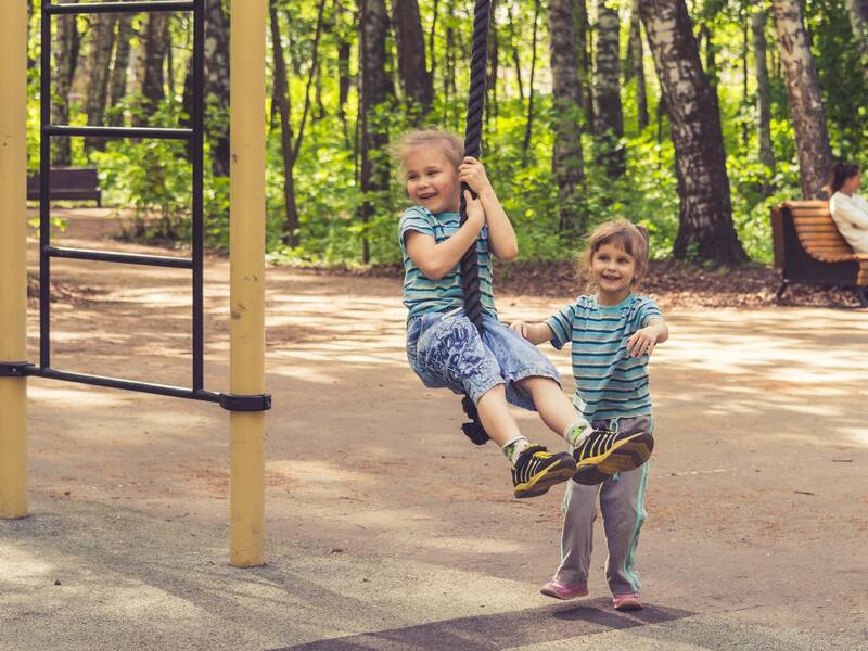 Zwei lachende Kinder spielen im Freien an einem Klettergerüst in einem bewaldeten Park.