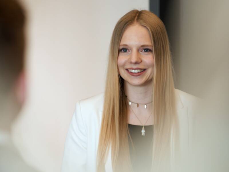 Portrait einer Frau. Sie trägt einen weißen Blazer, ein schwarzes Oberteil darunter und sie lächelt. Sie hat lange, glatte, blonde Haare. 