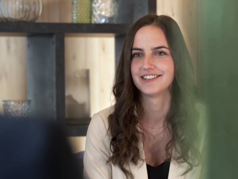 Portrait einer Frau mit dunklen, lockigen, schulterlangen Haaren. Sie lächelt leicht und trägt einen beigen Blazer. 