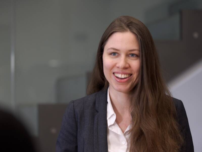 Portrait einer Frau mit welligen, langen, braunen Haaren. Sie trägt eine weiße Bluse und einen dunkelblauen Blazer. Ihr Mund ist leicht geöffnet. 