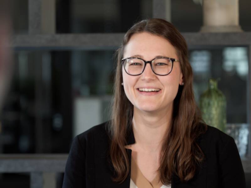 Portrait einer Frau mit einer schwarzen Brille und einem schwarzen Blazer. Sie hat braune. schulterlange Haare und hat auf beiden Seiten oben zwei Strähnen nach hinten gebunden. 