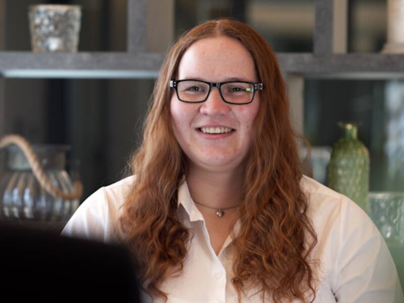 Portraitaufnahme einer Frau mit langen roten und lockigen Haaren. Sie trägt eine weiße Bluse, eine schwarze Brille und schaut die Person ihr gegenüber an. 