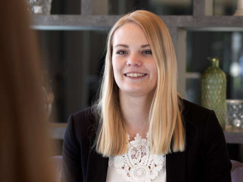 Eine Frau mit längeren blonden Haaren. Sie trägt ein weißes Oberteil und darüber einen schwarzen Blazer. Im Hintergrund ist ein Regal mit Deko und einer grünen Vase 