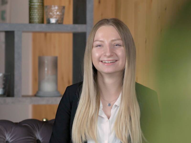 Portraitaufnahme eine Frau mit langen blonden Haaren. Ihr Mund ist geöffnet und sie lacht. Sie trägt einen schwarzen Blazer und eine weiße Bluse.