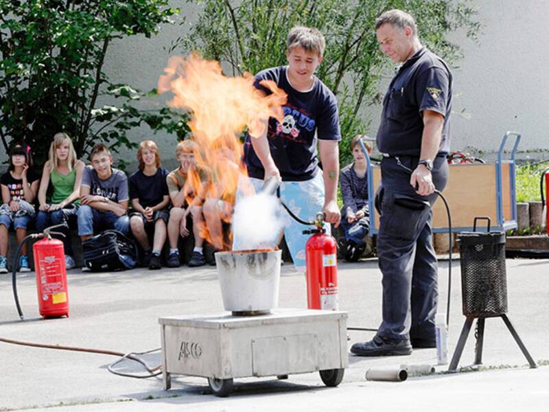 Zwei Männer stehen um einen großen Topf herum, aus dem ein großer Feuer herausragt. Der hintere Mann hat einen Feuerlöscher in der Hand. Im Hintergrund sitzen Jugendliche auf einer Bank und schauen den beiden Männern zu, die das Feuer machen und sich darum kümmern. 