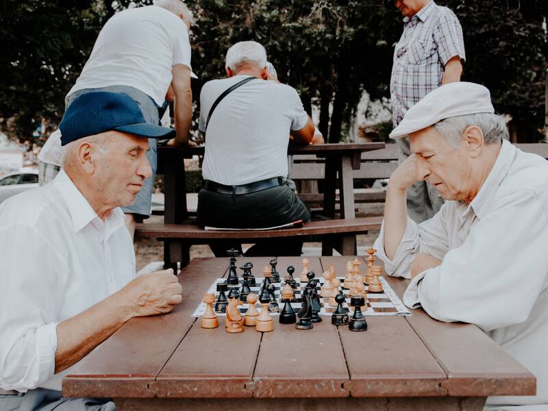 Zwei ältere Männer die an einem Tisch sitzen und Schach spielen. Beide schauen konzentriert auf das Spielbrett, das auf dem Tisch steht. Im Hintergrund sind drei weitere Männer erkennbar. Zwei davon stehen um einen weiteren Tisch und einer sitzt davor. 