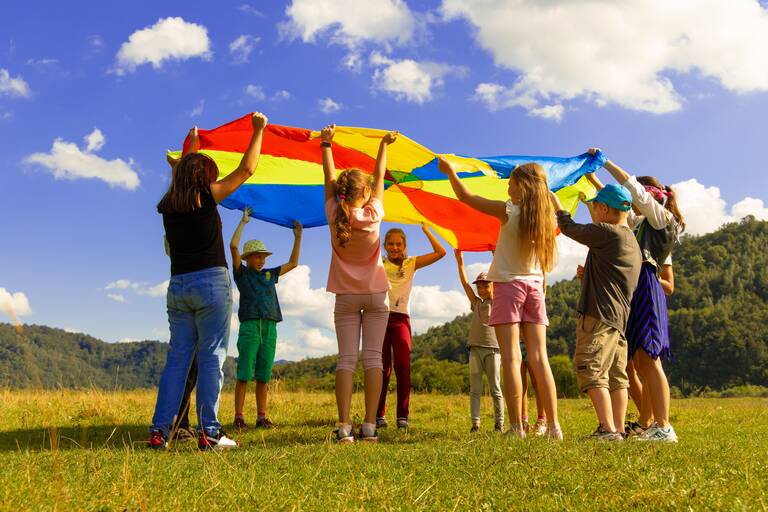 Kinder die in einem Kreis stehen. Sie halten nach oben ein buntes großes Tuch. Sie schauen sich unter dem Tuch durch an. 