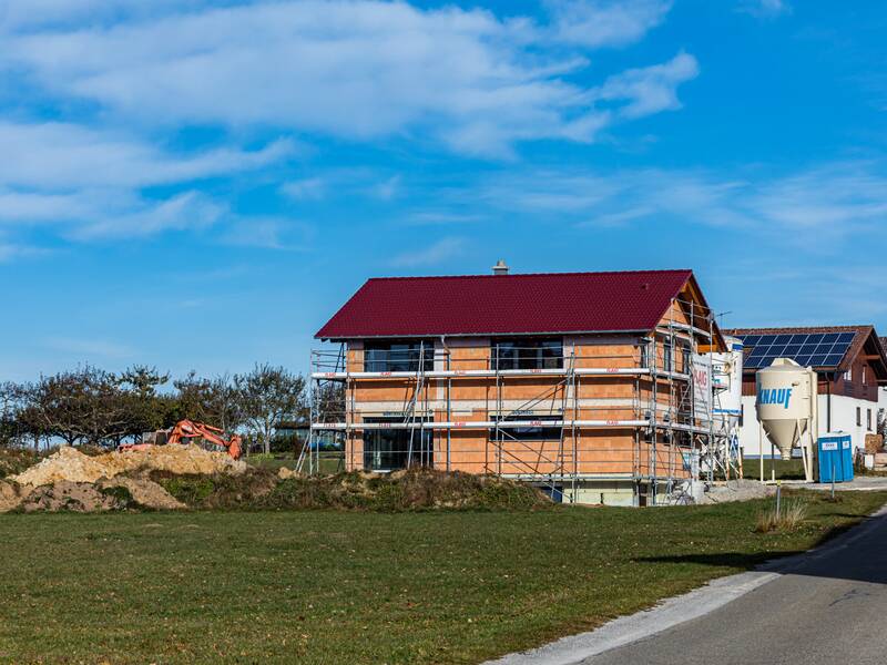 Neu gebautes Haus, das noch eingerüstet ist. Das Haus steht am Ende eines Wohngebiets auf einer grünen Wiese. Der Himmel ist blau.