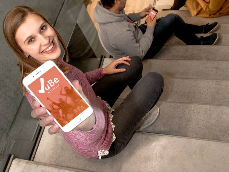 Ein Mädchen sitzt auf einer Betontreppe mit einem Mädchen im Vordergrund. Sie zeigt ein Handy die Treppe nach oben mit dem Bildschirm in die Kamera. im Hintergrund sitzen ebenfalls auf der Treppe ein weiterer Junge und ein Mädchen, die jeweils in ein Handy schauen.